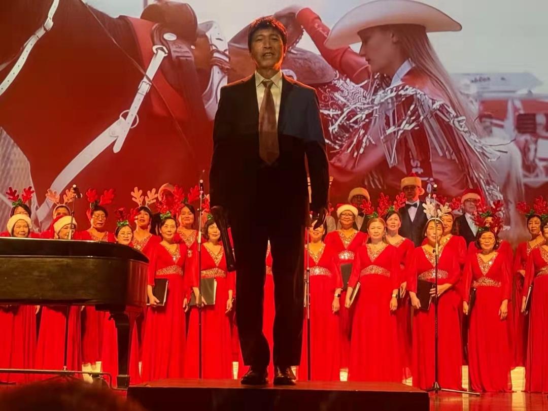 A person standing on a stage with a piano and a group of people in red dresses

Description automatically generated